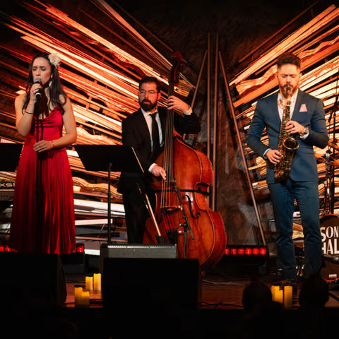 Foto do palco com uma cantora vestida de vermelho, um violoncelista de terno preto e um saxofonista de terno azul.